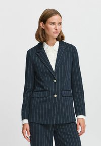 Tumesinise pinstripe ülikond, millel on kaks esikot ja kuldne nupp, omab kohandatud sobivust ja lõikega kraed, valmistatud tekstureeritud kangast.