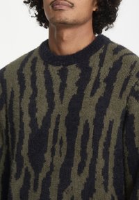 Pull en tricot vert olive avec des rayures abstraites bleu foncé. Il dispose d'un col rond et d'un tissu doux et texturé.