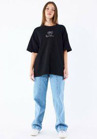 Camiseta negra de gran tamaño con un estampado gráfico en el frente, combinada con jeans rectos de mezclilla azul claro y zapatillas blancas.