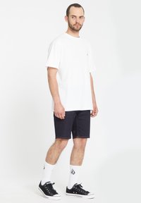 Witte T-shirt met korte mouwen gecombineerd met donkere denim shorts. Model draagt zwarte sneakers en witte sokken met een logo, staande op een lichte achtergrond.