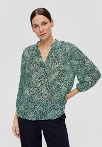 Groene blouse met een patroon, v-hals en geplooide details. Lichtgewicht, doorzichtig materiaal met een mix van groene en zwarte wervelingen. Lange mouwen.