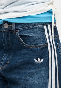 Τζιν παντελόνια σε σκούρο μπλε χρώμα με γκρι ανοιχτό τρεις ρίγες, με λογότυπο κεντημένο σε λευκό στο αριστερό μπούτι και αντίθετη ραφή.