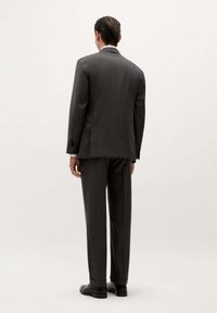 Hombre con el cabello peinado hacia atrás, vistiendo un traje gris oscuro y zapatos negros, de pie de espaldas con un fondo claro y sencillo.