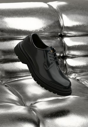 Black leather shoe with a glossy finish, rounded toe, thick black rubber sole, and contrasting laces. Narrow side cutout detail.