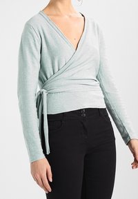 Jasnozielona bluzka w stylu kopertowym z miękkiego materiału, z głębokim dekoltem w kształcie V i długimi rękawami. Posiada wiązanie w talii i jest zestawiona z czarnymi spodniami.