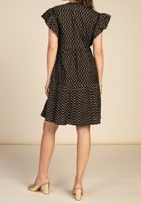 Schwarzes Kleid mit einem Zickzackmuster, fließendem Schnitt und rüschierten Ärmeln. Kombiniert mit goldenen Sandalen mit Blockabsatz. Rückansicht.