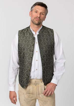 Mann trägt traditionelle bestickte beige Lederhosen, weißes Hemd und dunkle gemusterte Weste, steht vor einfachem Hintergrund.