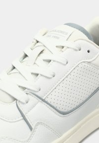 Bijele vezne tenisice sg gornjim dijelom od teksturirane kože, perforiranim bočnim panelom i svijetloplavim naglaskom blizu pete. Ravni gumeni potplat.