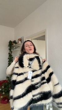 Bundová bunda s pruhovaným vzorom v krémovej a čiernej farbe. Má široký golier, pripevnenú visačku a mäkkú, nadýchanú textúru.