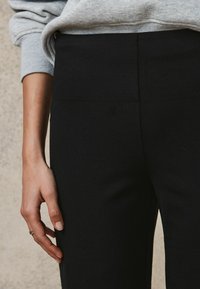 Czarne legginsy z wysokim stanem wykonane z elastycznego materiału. Dłoń oparta jest z boku, co podkreśla gładką fakturę i prosty design.