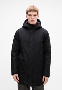 Jonge man in een zwarte winterjas met capuchon, voorzien van ritssluitingen aan de zijkanten en een ritszak op de linker mouw, staande tegen een witte achtergrond.
