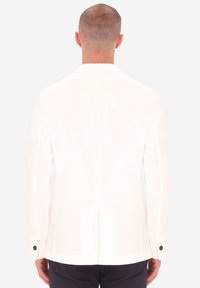 Uomo con capelli corti che indossa un blazer bianco e pantaloni scuri, in piedi di spalle contro uno sfondo chiaro e uniforme.