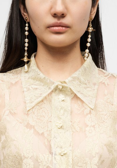 Διάφανη, ελαφριά κρεμ μπλούζα με φλοράλ δαντελένιο μοτίβο, με κλασικό γιακά, λεπτομέρειες από πέρλες και κουμπιά σε σχήμα τριαντάφυλλου. Μακριά, κρεμαστά σκουλαρίκια με πέρλες.