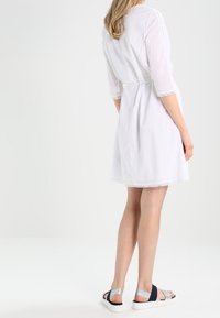 Robe blanche à manches trois-quarts, taille cintrée, bordure en dentelle aux extrémités des manches, associée à des sandales multicolores à talon bloc.