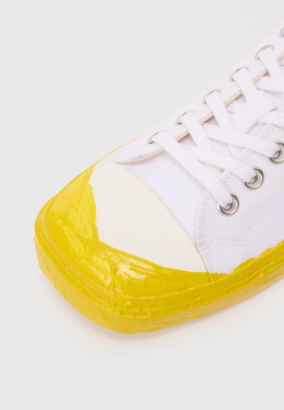White canvas shoe featuring a yellow rubber toe cap and sole, with yellow accents at the front. Rounded shape and flat design.