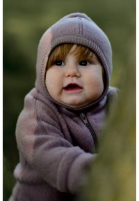 Otrok v vijoličasti flis obleki z zadrgo in kapuco. Tekstura je videti mehka in topla. Kapa ima rebrast rob, ozadje je zamegljeno.