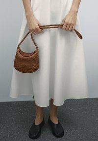 Brązowa pleciona torebka z zaokrąglonym dnem i plecionym uchwytem, w zestawieniu z białą sukienką i czarnymi mokasynami na neutralnym tle.