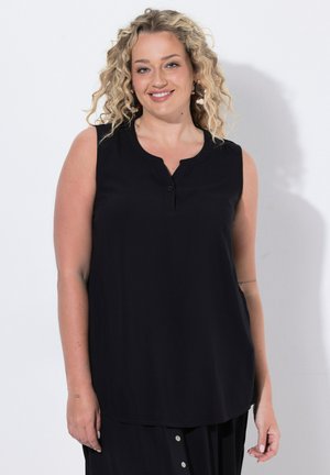 Femme souriante aux cheveux blonds bouclés portant un haut noir sans manches et une jupe noire, debout devant un fond blanc.