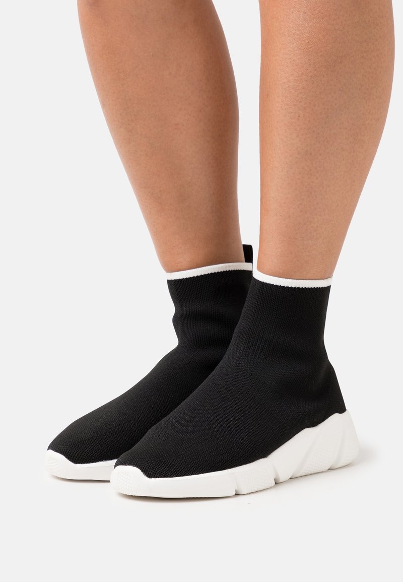 Jambes portant des baskets style chaussettes noires avec semelles et bordures blanches, debout sur un fond blanc uni.