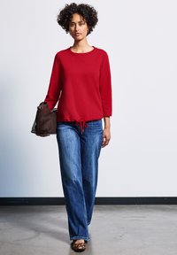 Röd sweatshirt med lång ärm och dragsko i nederkant, kombinerad med ljusblå vida jeans och en brun perforerad handväska.