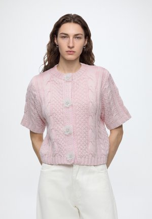 Kvinde med langt brunt hår iført en lyserød kortærmet strikket cardigan med blomsterformede knapper og hvide bukser, stående mod en hvid baggrund.