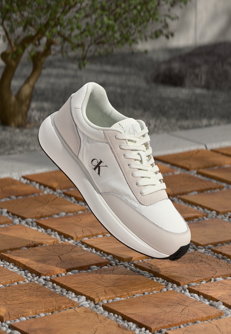 Sneaker bianco Calvin Klein con lacci, esposto su piastrelle di legno e piccoli ciottoli bianchi in un ambiente esterno con un albero sullo sfondo.