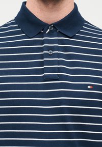 Πόλο μπλε ναυτικό με λευκές οριζόντιες ρίγες, με κλασικό γιακά και πατιλέτα με δύο κουμπιά. Μικρό κεντημένο λογότυπο στο στήθος.