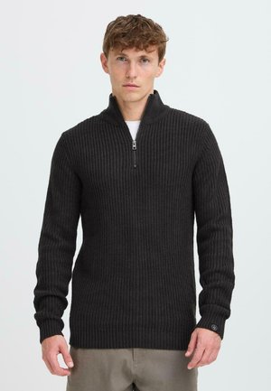Suéter de cuello con cremallera de color gris oscuro, con textura acanalada, mangas largas y un ajuste ligeramente holgado. Presenta un pequeño logotipo en el puño.