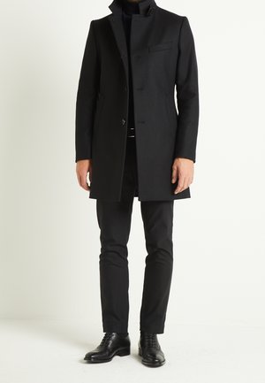 Homme portant un manteau noir boutonné à col montant, un pantalon noir et des chaussures habillées noires et polies, debout devant un fond clair uni.