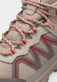 Μπεζ και γκρι πεζοπορικό αθλητικό παπούτσι με κόκκινες γεωμετρικές λεπτομέρειες, υφασμάτινο πλέγμα με υφή και μπεζ κορδόνια με κόκκινες βούλες δεμένα στην κορυφή.
