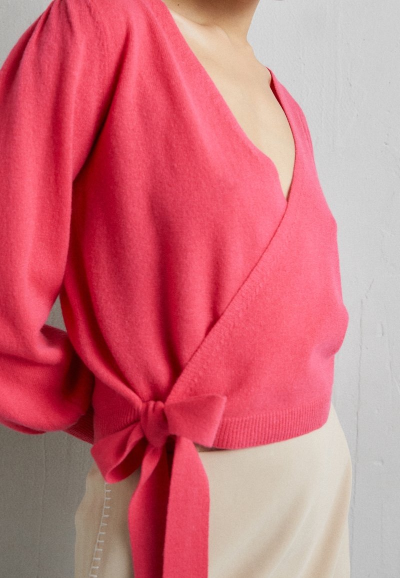 Pull cache-cœur rose en tissu doux, avec un col en V profond, des manches longues et un détail noué à la taille. Texture lisse et coupe ajustée.