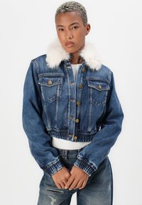 Chaqueta de mezclilla recortada en azul oscuro con cuello de peluche sintético, botones de metal, dos bolsillos en el pecho y puños elásticos. Llevada sobre una blusa blanca.