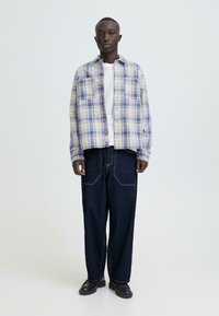 Licht geruit overhemd in beige en blauw, met borstzakken en knopen, gecombineerd met donkere denim broek en zwarte schoenen.