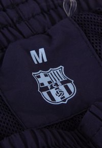 Middelgrote donkere stoffen label met het FC Barcelona-logo, dat een kruis, strepen en een bal toont, op sportkleding.