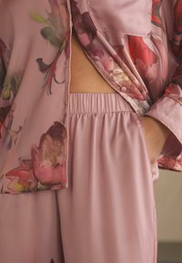 Ružový saténový loungewear set obsahujúci top s kvetinovou potlačou a gombíkovým zapínaním a nohavice s elastickým pásom a voľným strihom.