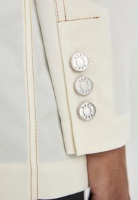 Manche de veste couleur crème avec trois boutons logo argentés, couture contrastante et texture lisse. Le tissu semble résistant et bien taillé.