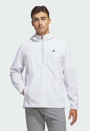 Mladý muž v bílé mikině na zip Adidas s rukama v kapsách stojí před jednobarevným světlým pozadím.