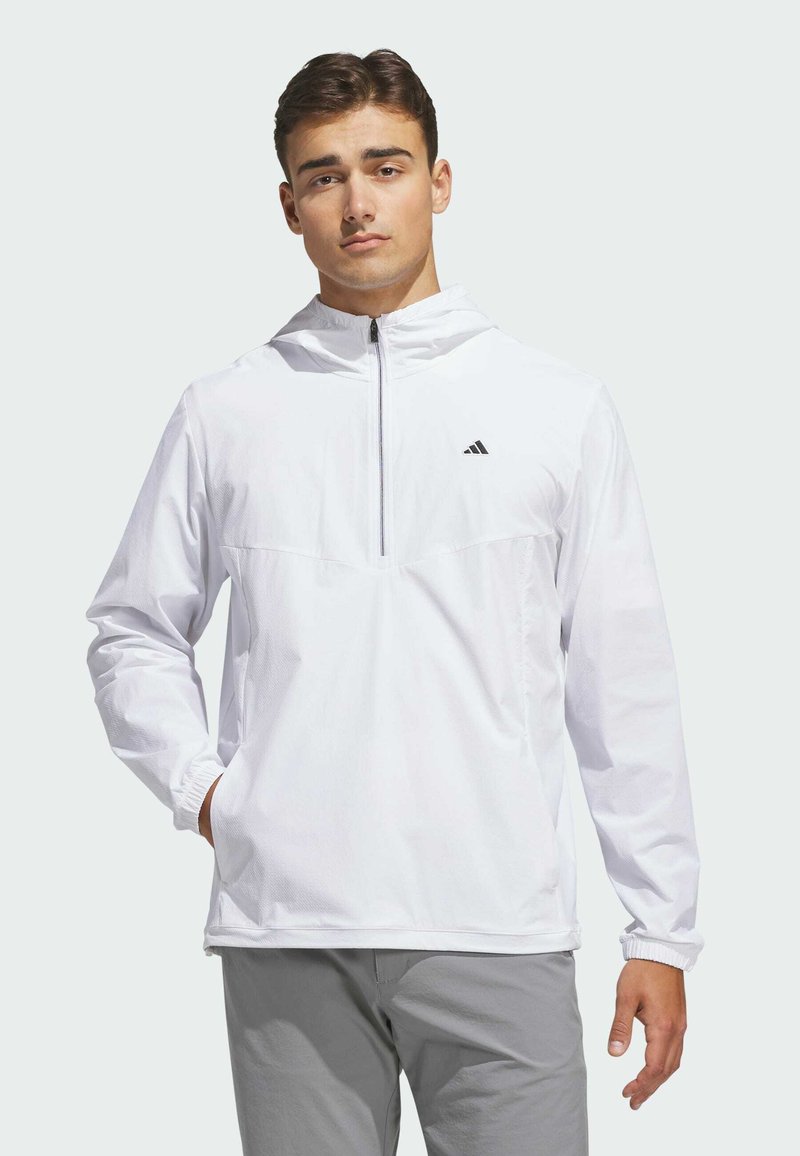 Jeune homme portant un sweat à capuche zippé blanc Adidas, les mains dans les poches, debout devant un fond clair uni.