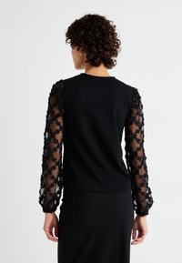 Svart topp med långa genomskinliga spetsärmar prydda med blomdekorationer. Slät tygkropp, rund halsringning och figurnära silhuett.