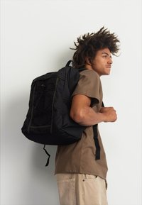 Μαύρο σακίδιο από ύφασμα με κίτρινες ραφές, ορθογώνιο σχήμα, μπροστινή τσέπη και ρυθμιζόμενες ιμάντες, που φοριέται από άτομο που κοιτάζει πλάγια.