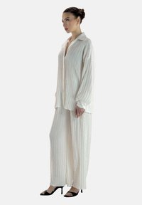 Ensemble deux pièces en tissu blanc froissé comprenant une chemise boutonnée à manches longues et un pantalon à jambes larges, tous deux avec une texture à motif vertical.