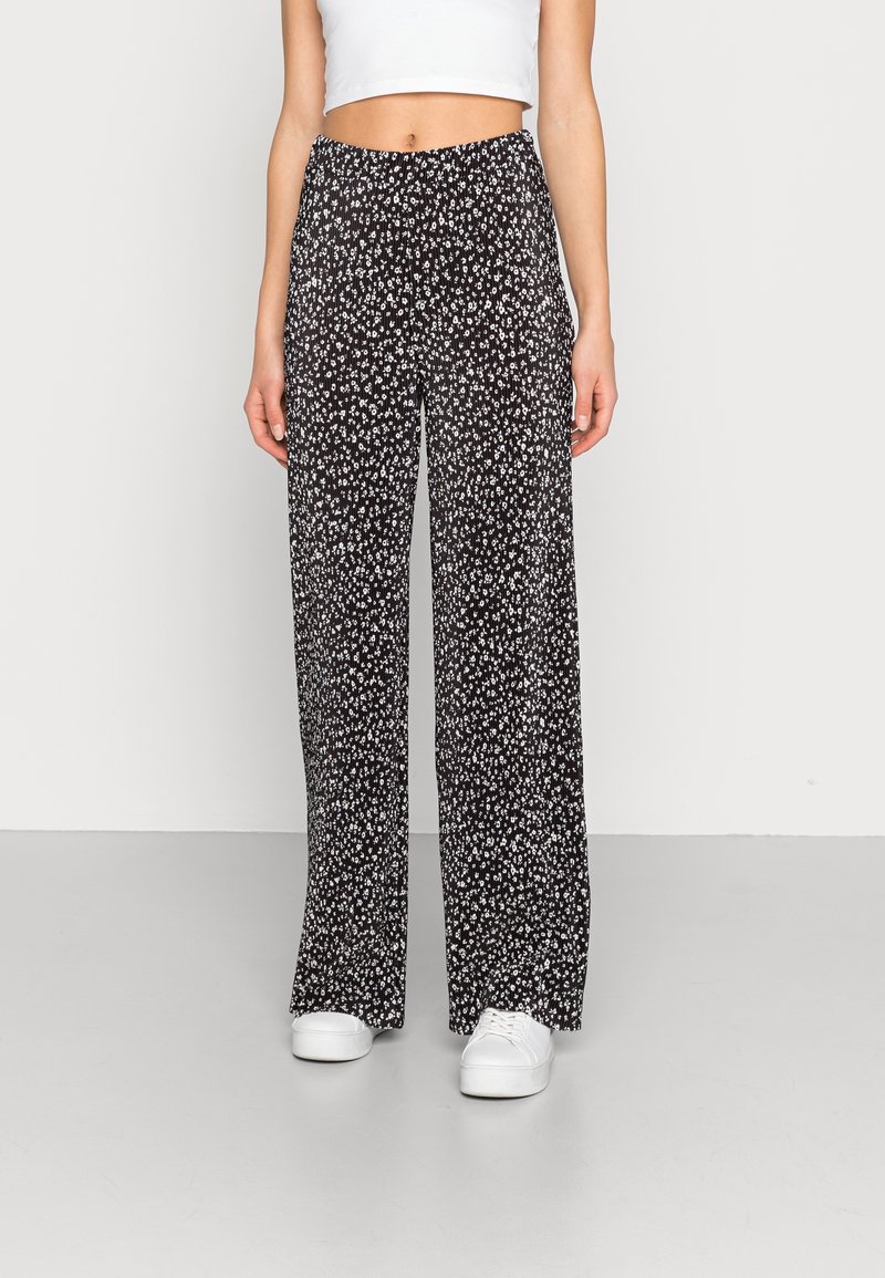 Pantalon noir à jambes larges avec un petit motif floral blanc, en tissu léger, doté d'une taille élastique. Assorti à des baskets blanches.