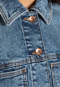 Close-up of blue denim jacket front with copper buttons and visible orange stitching around collar and pockets.