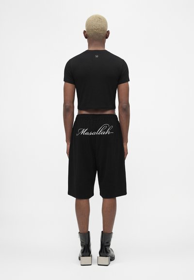 Άτομο με κοντά ξανθά μαλλιά φοράει μαύρο crop top, μαύρα σορτς με τη λέξη "Masallah" στην πλάτη και μαύρες μποτάκια με τακούνι, στέκεται με την πλάτη γυρισμένη.