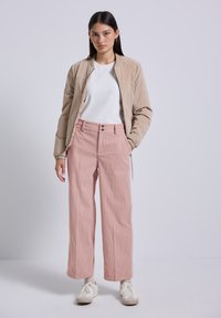 Giovane donna in piedi, che indossa una giacca bomber beige, una maglietta bianca, pantaloni in velluto a coste rosa a gamba larga e sneakers bianche, su uno sfondo semplice.