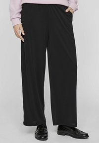 Crne široke hlače izrađene od glatkog materijala s elastičnom trening. Model nosi sjajne crne cipele s detaljima.