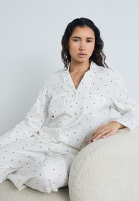 White pajama set with red heart patterns. Made of lightweight fabric, featuring a button-up top and loose-fitting pants. Textured background.