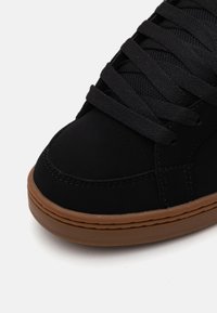 Schwarze Sneakers mit einem glatten, wildlederähnlichen Obermaterial, ausgestattet mit flachen schwarzen Schnürsenkeln und einer beige Gummisohle. Dezente Perforationen schmücken den Zehenbereich.