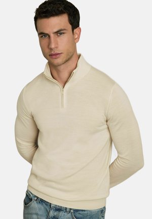 Beige ritsvest gemaakt van zacht materiaal. Heeft een hoge kraag, een aansluitende pasvorm en geribbelde manchetten. Gdragend met blauwe jeans.