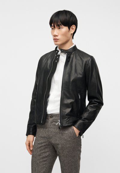 Νέος άνδρας με μαύρο δερμάτινο μπουφάν πάνω από λευκό πουκάμισο με κουμπιά και γκρι υφασμάτινη παντελόνα, στέκεται με το ένα χέρι στην τσέπη.